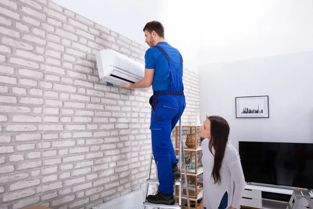 HVAC installer on a ladder mounting an indoor AC unit while a female customer watches.