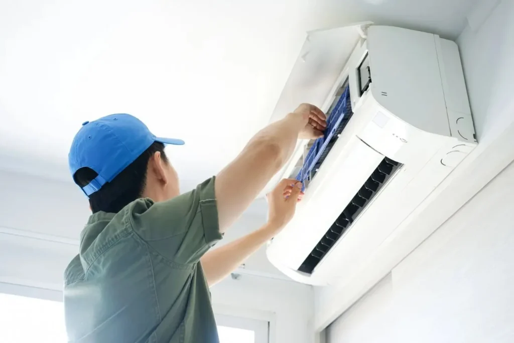 A technician in a blue cap is performing repair or maintenance on a wall-mounted AC unit.
